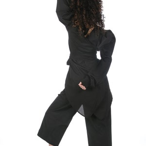 May include: A black outfit consisting of a wrap top with long sleeves, wide-leg pants, and white sneakers. The top has a unique asymmetrical design. The model's curly hair is visible.