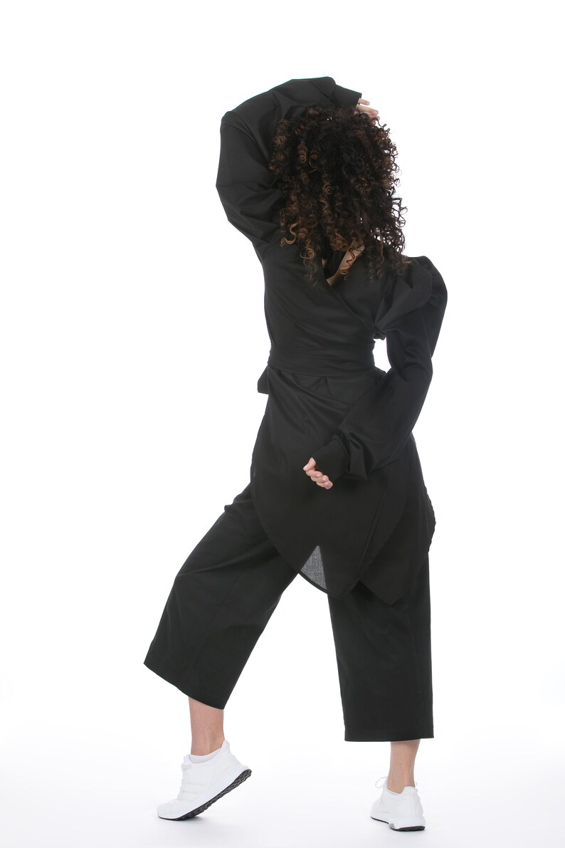 May include: A black outfit consisting of a wrap top with long sleeves, wide-leg pants, and white sneakers. The top has a unique asymmetrical design. The model's curly hair is visible.