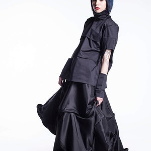 May include: A woman wearing a black top with a high collar and a black skirt with multiple layers. The skirt is flowing and the woman is looking down.