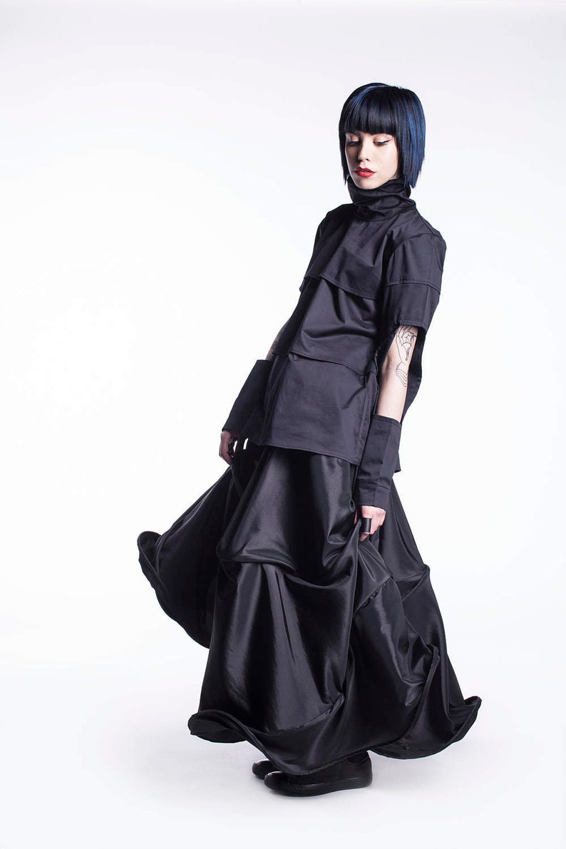 May include: A woman wearing a black top with a high collar and a black skirt with multiple layers. The skirt is flowing and the woman is looking down.