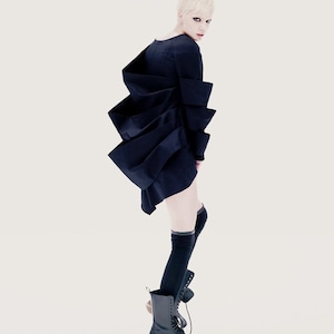 May include: A woman with short blonde hair wearing a black dress with a unique layered design. She is also wearing black knee-high socks and black lace-up boots.
