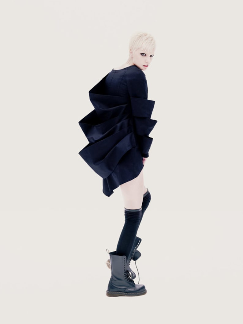 May include: A woman with short blonde hair wearing a black dress with a unique layered design. She is also wearing black knee-high socks and black lace-up boots.