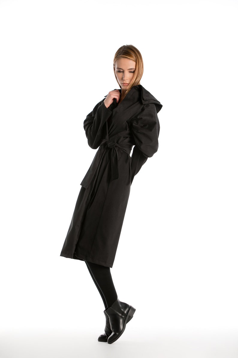 May include: A woman wearing a black hooded coat with a belt. She is standing on a white background.