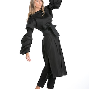 Op de afbeelding: Een zwarte lange mouwen jurk met een getailleerde taille en grote, geplooide mouwen. De jurk heeft een split aan de voorkant en wordt gecombineerd met een zwarte broek en zwarte schoenen.
