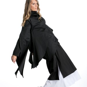 May include: A woman wearing a black, asymmetrical shirt with a high collar and a black skirt with a white underskirt. She is wearing black shoes with a yellow sole.