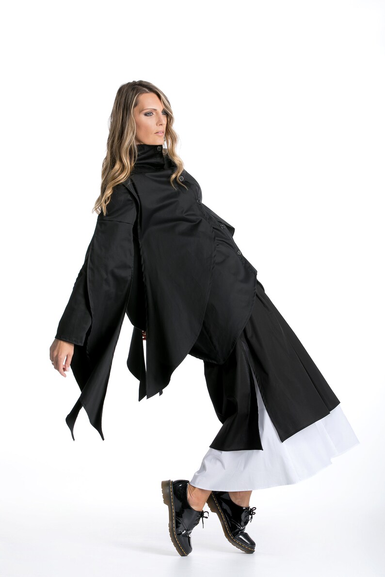 May include: A woman wearing a black, asymmetrical shirt with a high collar and a black skirt with a white underskirt. She is wearing black shoes with a yellow sole.