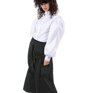 May include: A woman wearing a white button-down shirt with large, puffy sleeves and a black skirt. She is wearing a black visor and white sneakers.