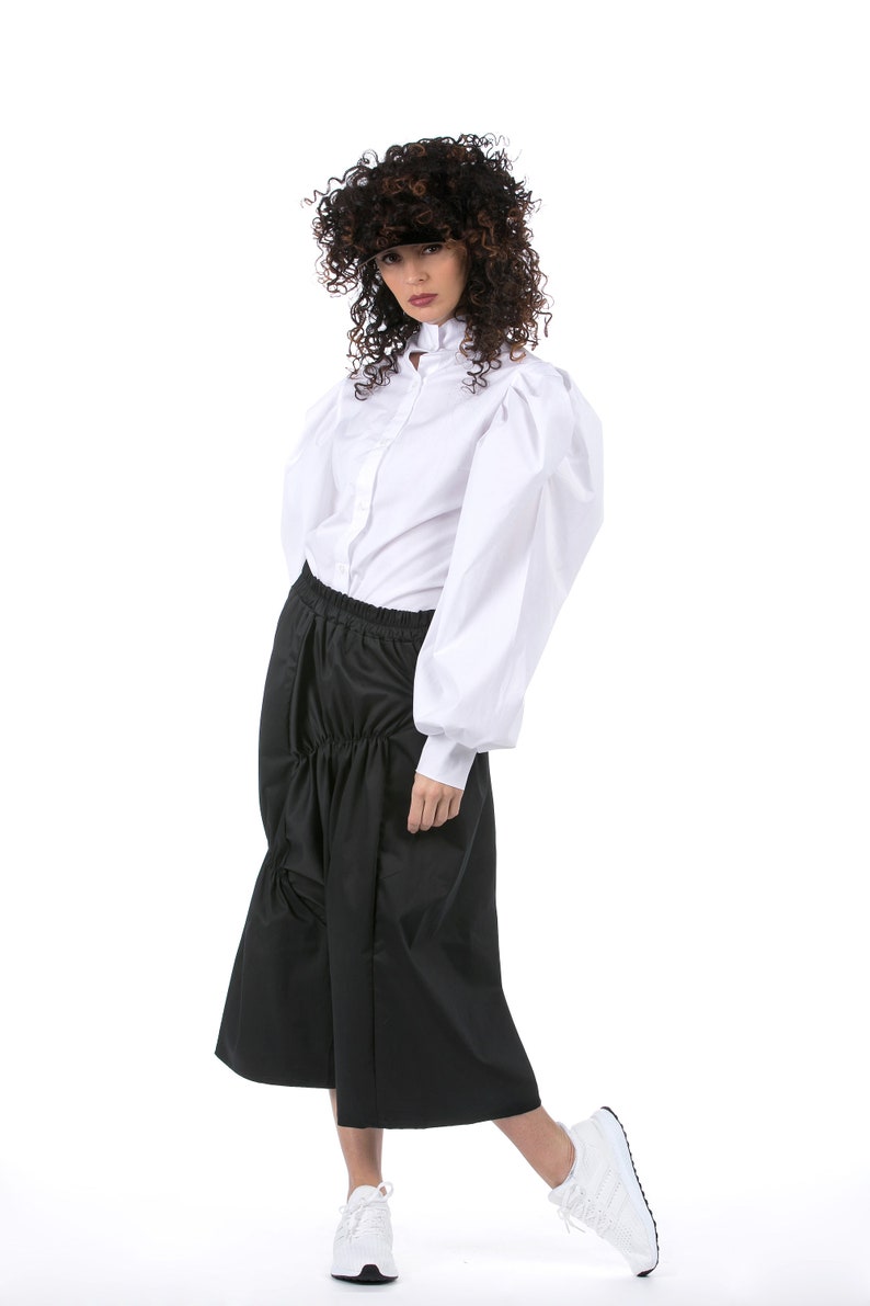 May include: A woman wearing a white button-down shirt with large, puffy sleeves and a black skirt. She is wearing a black visor and white sneakers.