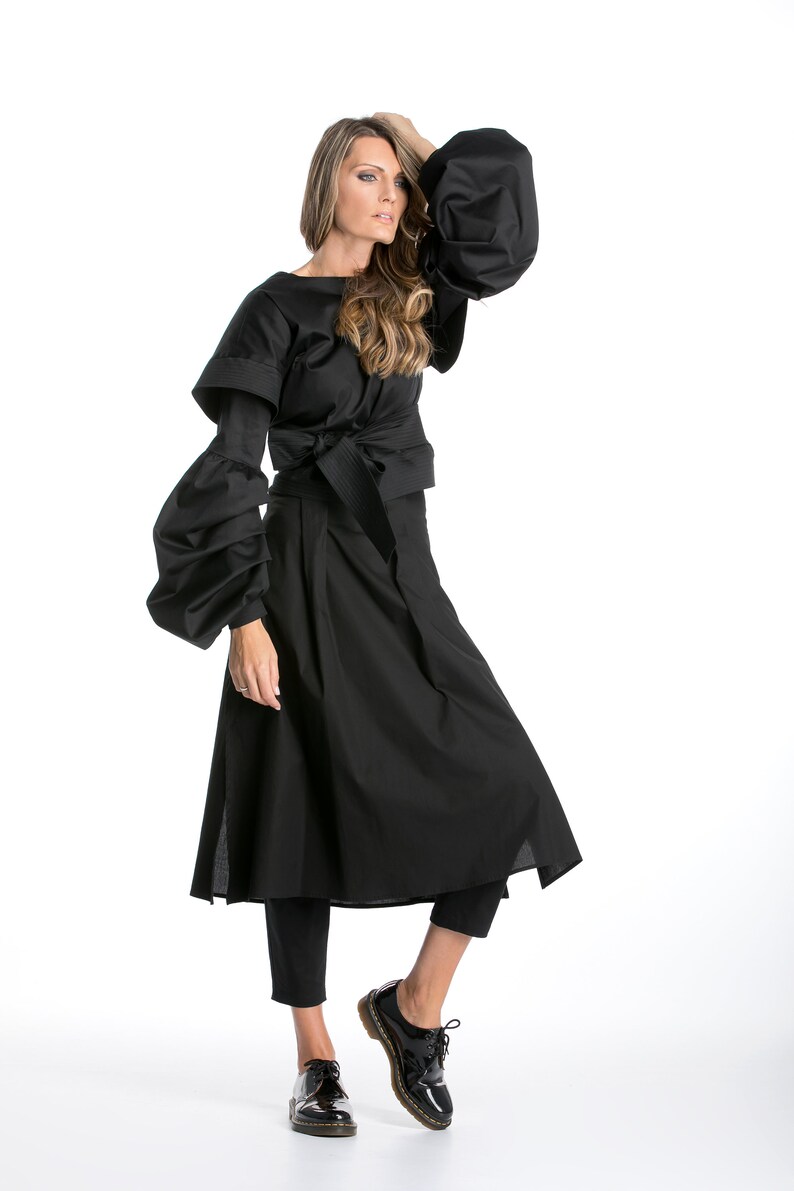 Op de afbeelding: Een zwarte jurk met een lange rok en een getailleerd bovenstuk. De jurk heeft grote, pofmouwen en een zwarte riem. Het model draagt een zwarte broek en zwarte schoenen.