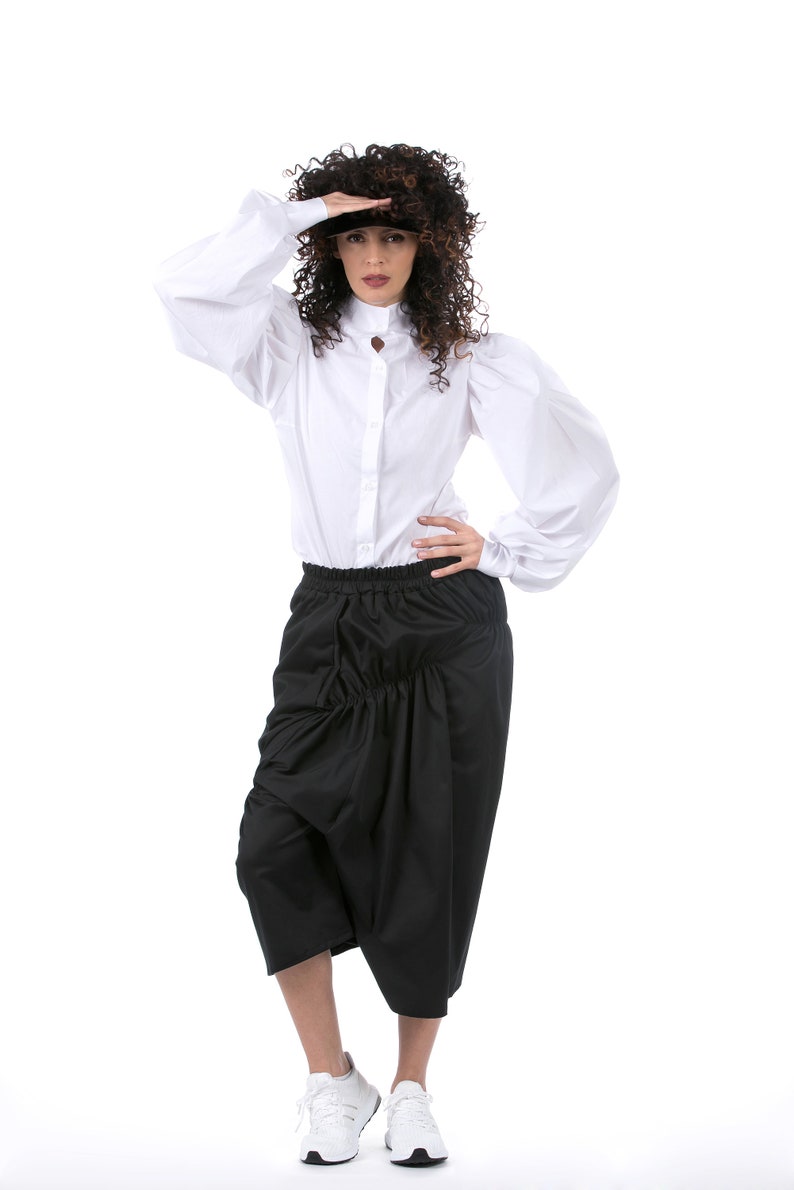 May include: A woman wearing a white button-down shirt with puffed sleeves and a black gathered skirt. She is wearing white sneakers.