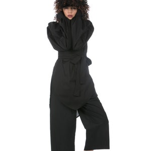 May include: A black jumpsuit with a wrap-style top and wide-leg pants. The outfit is cinched at the waist with a matching belt. The person is wearing white sneakers. The model has curly brown hair.