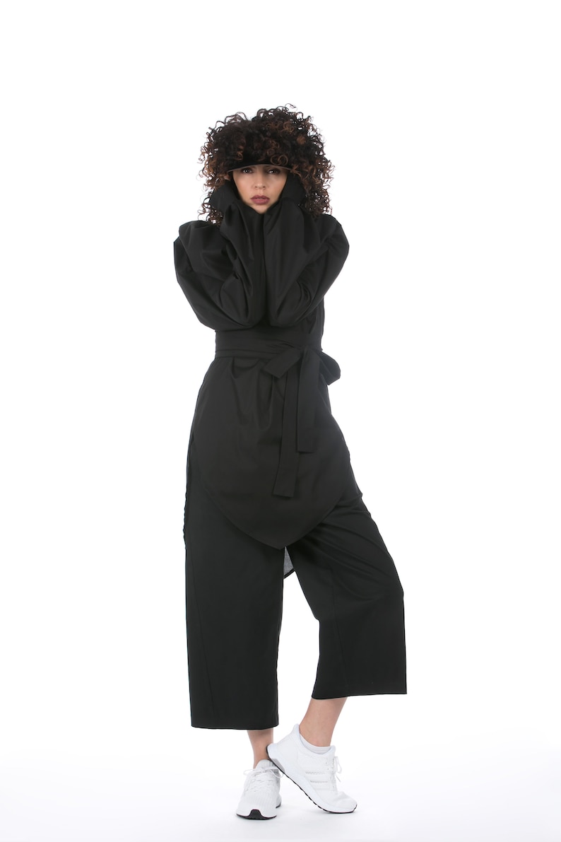 May include: A black jumpsuit with a wrap-style top and wide-leg pants. The outfit is cinched at the waist with a matching belt. The person is wearing white sneakers. The model has curly brown hair.