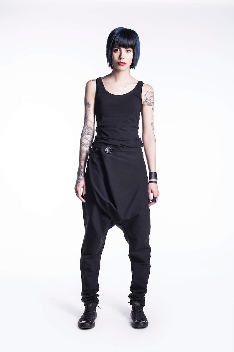 May include: A woman wearing a black tank top and black pants with a unique design. She has short, dark blue hair and tattoos on her arms.