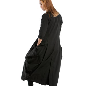 K&ouml;nnte beinhalten: Eine Frau in einem langen schwarzen Kleid mit gerafftem Rock und Taschen. Sie tr&auml;gt schwarze Stiefeletten und hat lange braune Haare.