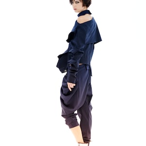 May include: A woman wearing a dark blue, asymmetrical top with a high neckline and a pair of dark blue, loose-fitting pants. She is wearing white sneakers with brown laces.