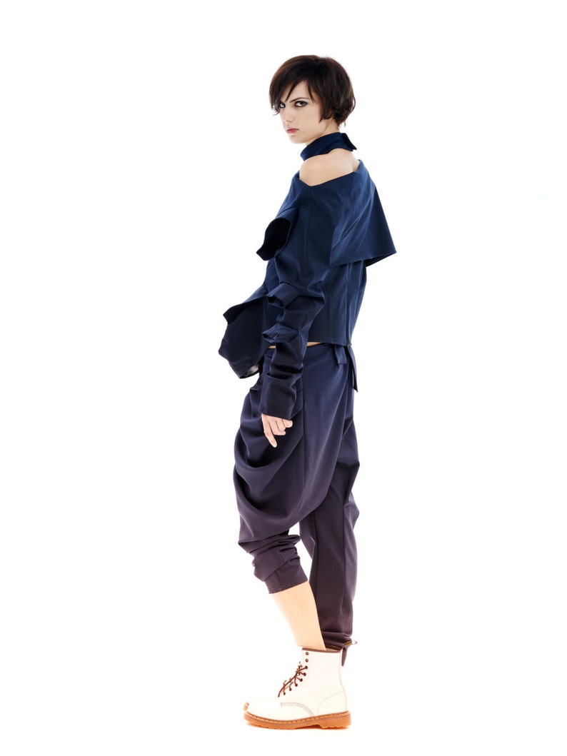 May include: A woman wearing a dark blue, asymmetrical top with a high neckline and a pair of dark blue, loose-fitting pants. She is wearing white sneakers with brown laces.