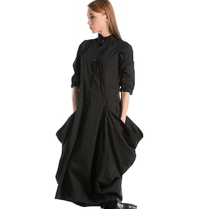 K&ouml;nnte beinhalten: Ein schwarzes, lang&auml;rmliges Kleid mit Knopfleiste und gro&szlig;en Taschen. Das Kleid hat einen flie&szlig;enden, asymmetrischen Saum und wird mit schwarzen Stiefeletten getragen.