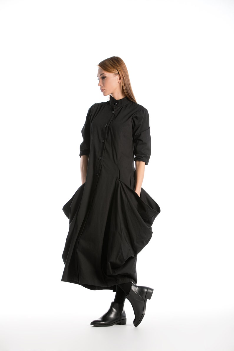 K&ouml;nnte beinhalten: Ein schwarzes, lang&auml;rmliges Kleid mit Knopfleiste und gro&szlig;en Taschen. Das Kleid hat einen flie&szlig;enden, asymmetrischen Saum und wird mit schwarzen Stiefeletten getragen.