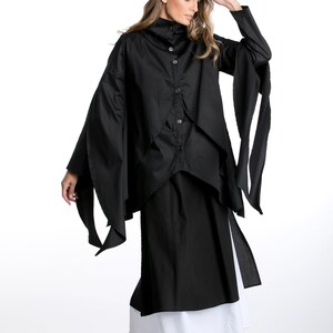 May include: A black long-sleeved shirt with a high collar and a layered design. The shirt has long, flowing sleeves that tie at the wrists. The model is wearing a black skirt and white underskirt.