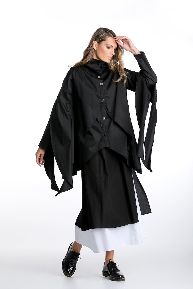 May include: A black long-sleeved shirt with a high collar and a layered design. The shirt has long, flowing sleeves that tie at the wrists. The model is wearing a black skirt and white underskirt.