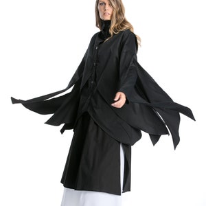 May include: A black long-sleeved shirt with a draped design and a white underskirt. The shirt has a button-up front and long, flowing sleeves. The model is wearing black shoes.