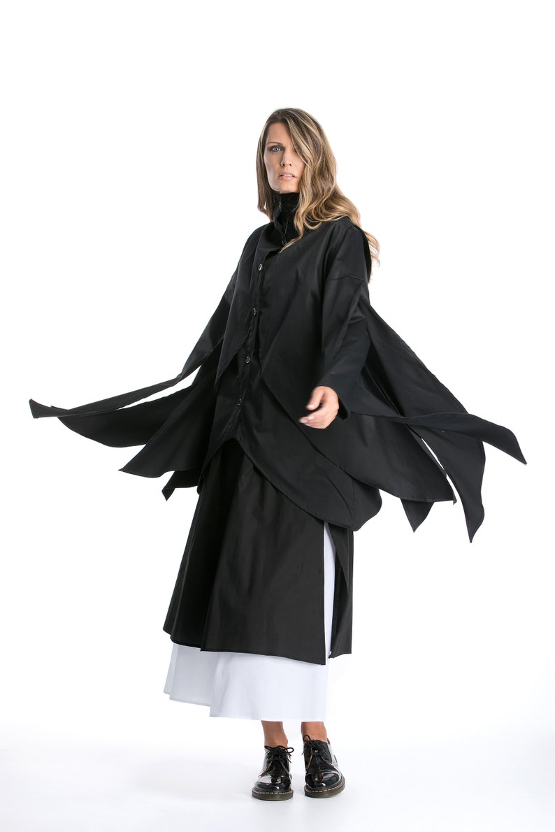 May include: A black long-sleeved shirt with a draped design and a white underskirt. The shirt has a button-up front and long, flowing sleeves. The model is wearing black shoes.