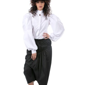 May include: A white button-up shirt with large, puffy sleeves and a black skirt with a gathered waist. The model is wearing white sneakers and a brown visor.