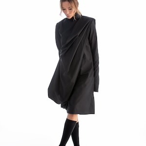 May include: A black long-sleeved dress with a draped design. The dress is worn by a person with blonde hair and black socks and shoes.