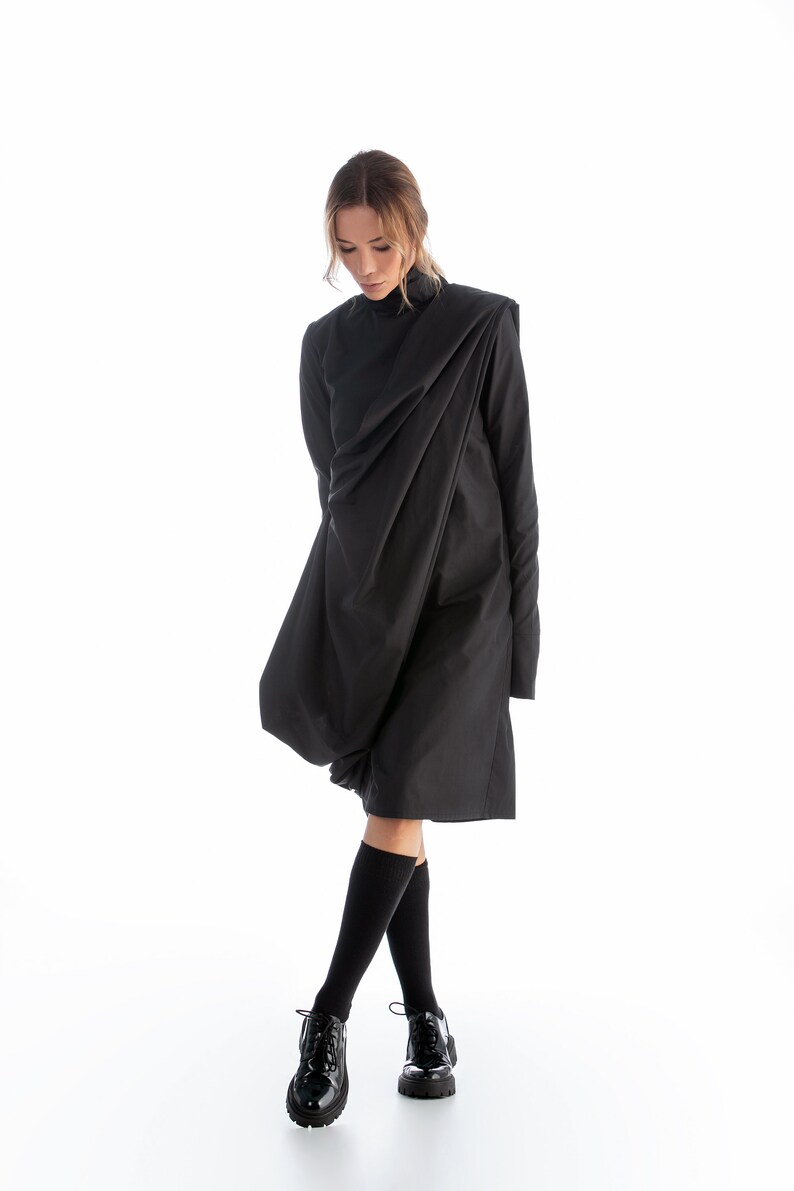 May include: A black long-sleeved dress with a draped design. The dress is worn by a person with blonde hair and black socks and shoes.