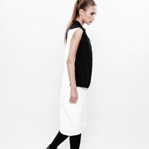 May include: A woman wearing a black and white sleeveless dress with black tights and black ankle boots. She is standing on a white background.