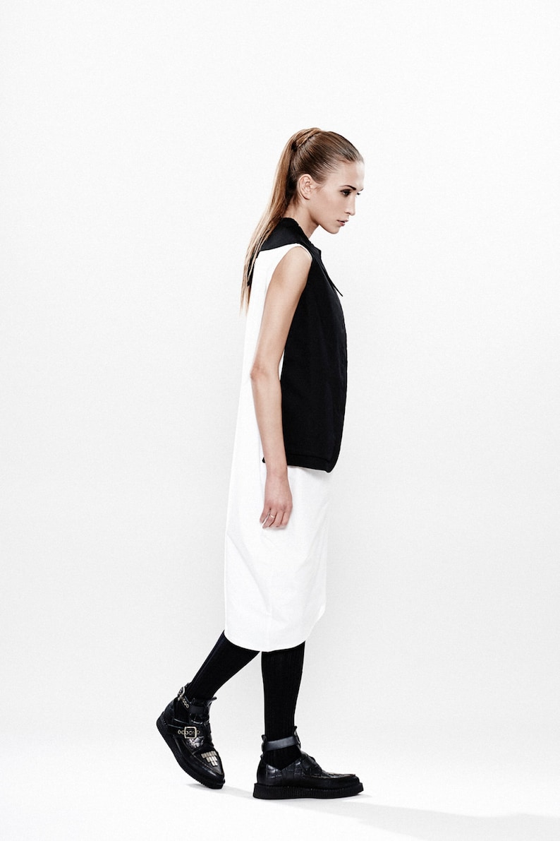 May include: A woman wearing a black and white sleeveless dress with black tights and black ankle boots. She is standing on a white background.
