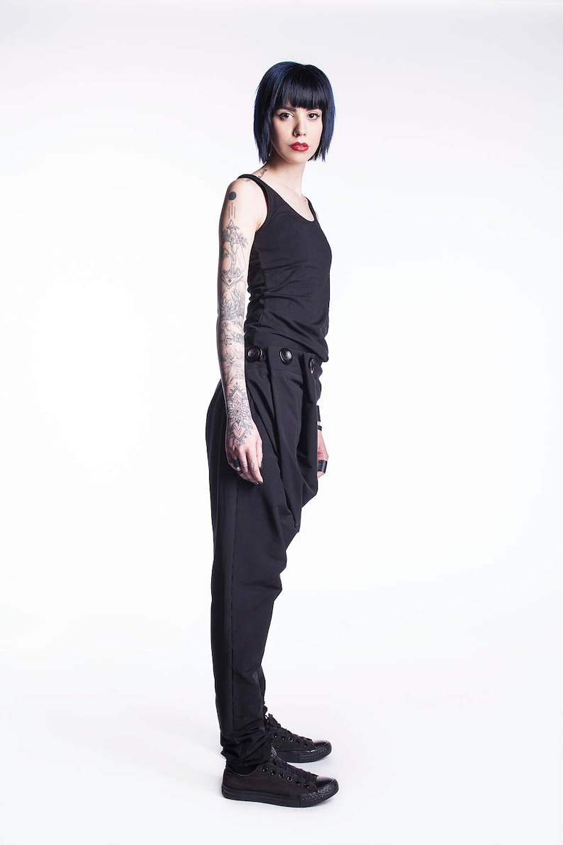 May include: A woman with short blue hair is wearing a black tank top and black pants with a unique design. She is wearing black sneakers and has tattoos on her arms.