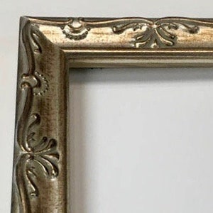 Pu&ograve; includere: Primo piano di una cornice per foto argentata con un design ornamentale. La cornice ha un bordo rialzato e una finitura liscia e lucida.