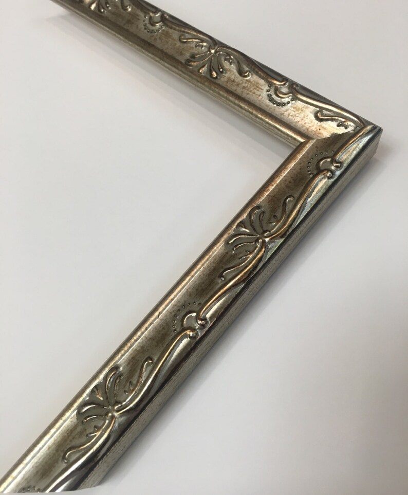 May include: A silver picture frame with an ornate design. The frame has a raised, textured border with a floral pattern.