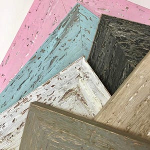 May include: Four wooden picture frames in various colours, including pink, blue, white, and grey. The frames are stacked on top of each other, with the pink frame on top and the grey frame on the bottom.