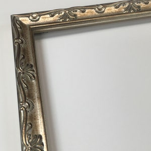 May include: A silver picture frame with an ornate design. The frame has a raised, textured border with a floral pattern.