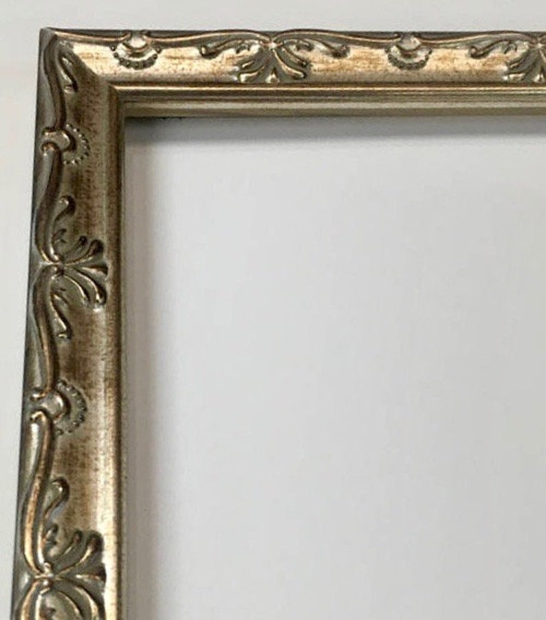 Pu&ograve; includere: Primo piano di una cornice per foto argentata con un design ornamentale. La cornice ha un bordo rialzato e una finitura liscia e lucida.