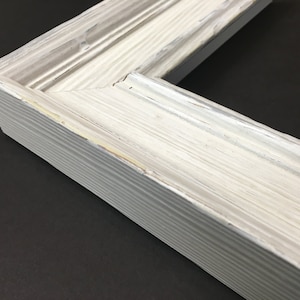 May include: Close-up of a white wood picture frame with a distressed finish. The frame has a raised edge and a simple design.