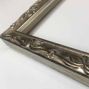 May include: A close-up of a silver picture frame with an ornate, detailed design. The frame has a slightly distressed finish and a subtle gold undertone.