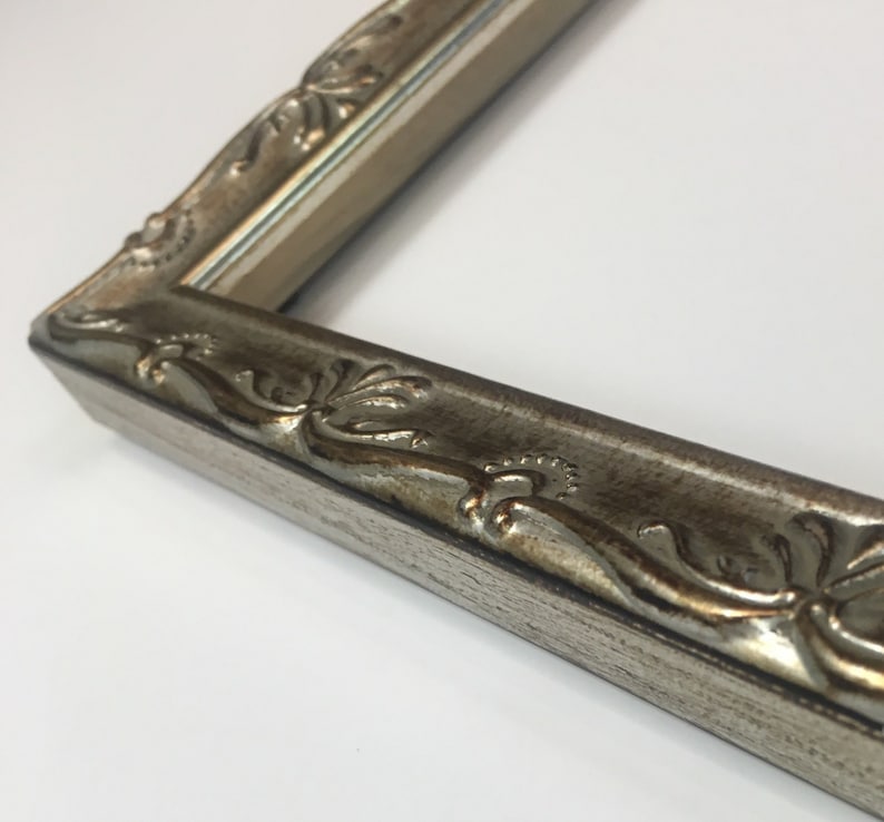 May include: A close-up of a silver picture frame with an ornate, detailed design. The frame has a slightly distressed finish and a subtle gold undertone.