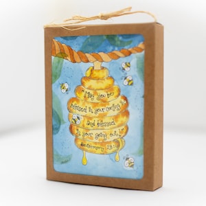 May include: A watercolour illustration of a beehive with the text "May you be blessed in your coming in and blessed in your going out!" on a card inside a brown box. The image includes bees and a rope. The card is tied with twine.
