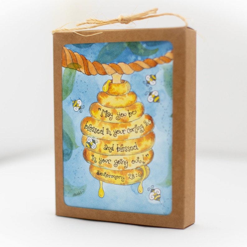 May include: A watercolour illustration of a beehive with the text "May you be blessed in your coming in and blessed in your going out!" on a card inside a brown box. The image includes bees and a rope. The card is tied with twine.