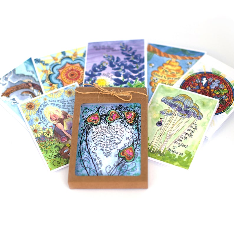 May include: A selection of illustrated greeting cards with watercolour designs. The cards depict flowers, mushrooms, and abstract patterns. A brown cardboard box with a window displays a card with a floral design and handwritten text. The cards are tied with twine.
