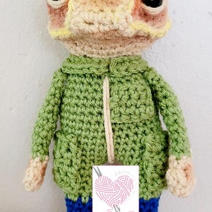 Puede incluir: Rana de crochet con una chaqueta verde y botas azules. La rana tiene una cabeza amarilla y naranja y ojos negros. La rana está de pie sobre una superficie estampada.