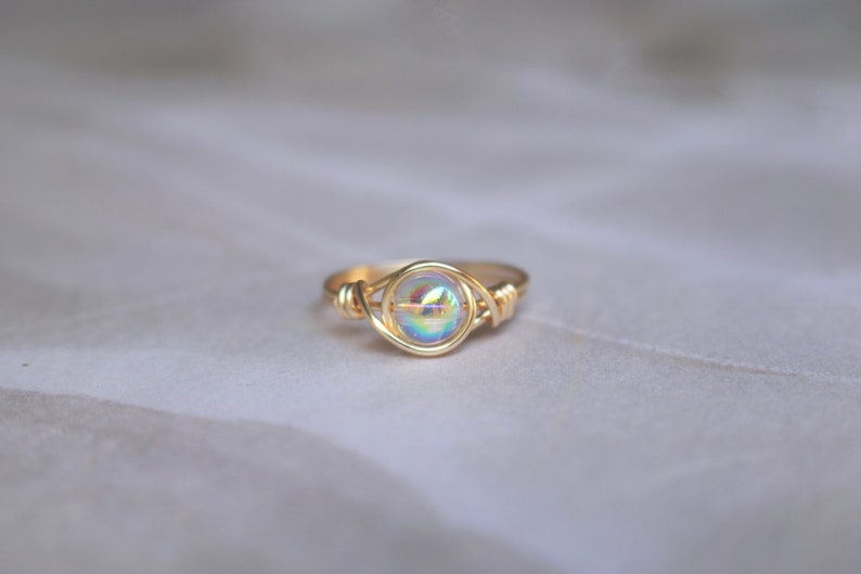 Pu&ograve; includere: Un anello avvolto in filo d'oro con una perla rotonda iridescente al centro.