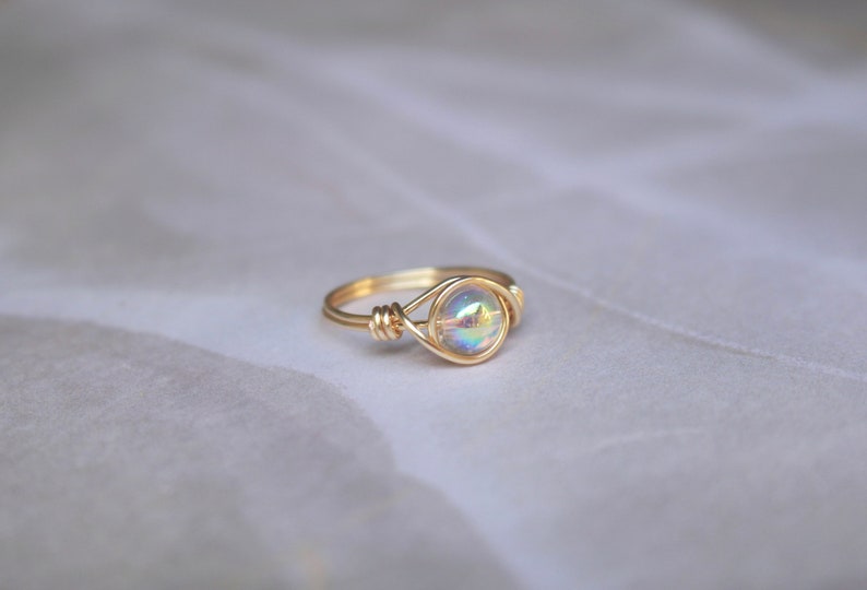 Pu&ograve; includere: Un anello in filo d'oro con una pietra rotonda iridescente.