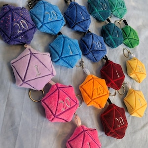 Könnte beinhalten: Eine Sammlung von weichen, mehrfarbigen zwanzigseitigen Würfel-Schlüsselanhängern in verschiedenen Farben wie Lila, Blau, Pink, Orange, Rot, Gelb und Grün. Jeder Stoffwürfel hat gestickte geometrische Linien und Zahlen wie 1 oder 20.