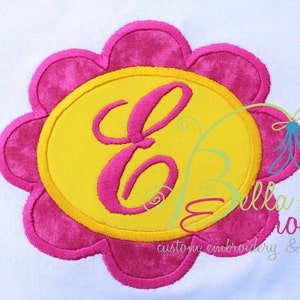 Puede incluir: Una flor rosa con un centro amarillo y la letra "E" bordada en rosa en su interior. La flor está rodeada de un círculo amarillo con un contorno rosa. El texto "Bella Bleu Bordado" y "bordado personalizado & diseños" es visible debajo de la flor.