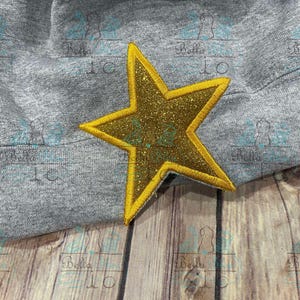 May include: A gold glitter star with a yellow outline, sewn onto a gray fabric background.