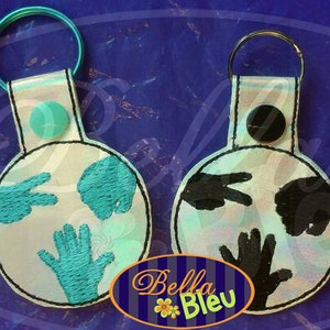 Puede incluir: Dos llaveros blancos con un diseño redondo e iridiscente. Un llavero presenta un gesto de mano azul, y el otro llavero presenta un gesto de mano negro.